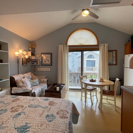 Cozy beach-themed studio with a bed, sofa, dining table, and large window overlooking a balcony.