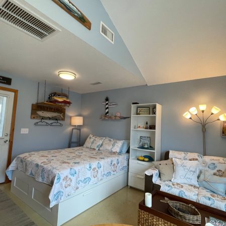 Cozy blue-themed bedroom with a bed, sofa, shelves, and nautical decor; natural light enters from a window and door.