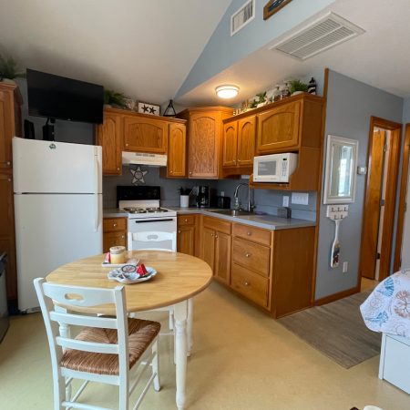 Small kitchen with wooden cabinets, white appliances, round dining table, and part of a bed in a compact studio space.
