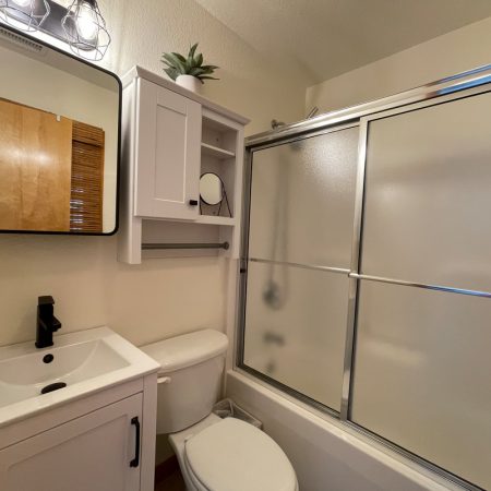 Small bathroom with white sink, toilet, cabinet, mirror, and a bathtub with sliding frosted glass doors.