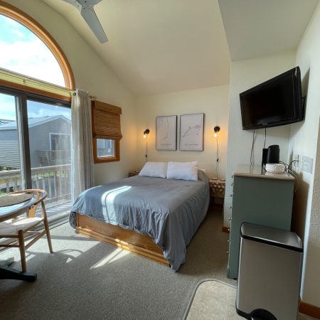 A small bedroom with a double bed, wall art, TV, table and chairs, large window, and natural light filling the space.