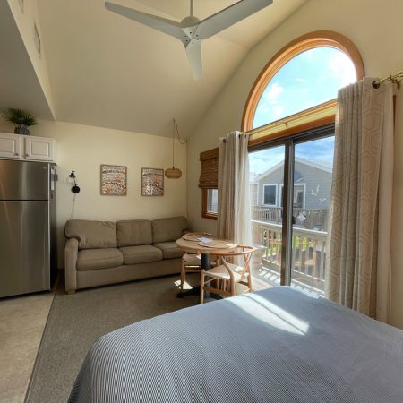 A cozy studio with a sofa, small dining table, kitchenette, and large window with sunlight streaming inside.