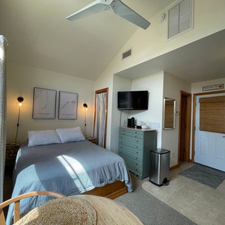 Compact studio with a neatly made bed, wall art, TV, kitchenette, and natural light from a window.