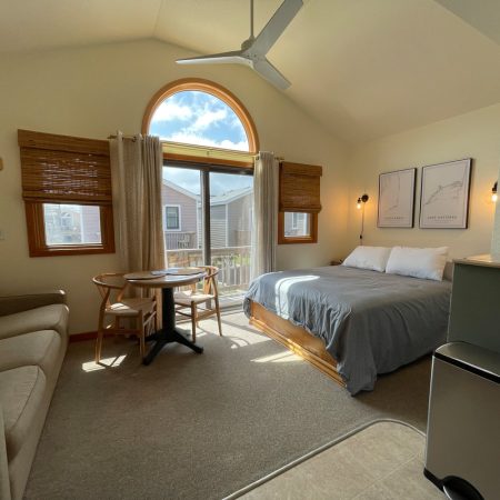 Sunny bedroom with a bed, sofa, round table, large window, wall art, TV, and mini fridge.