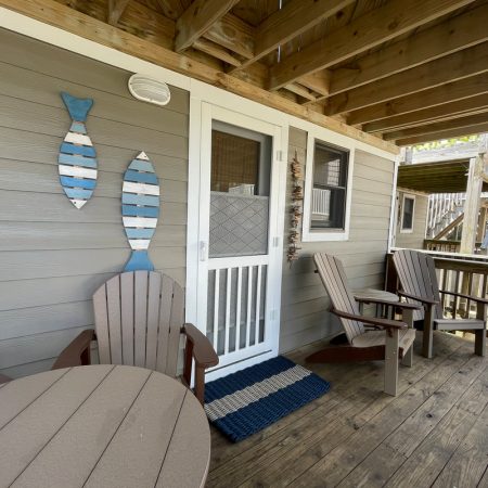 Wooden porch with two chairs, a round table, striped rug, and fish-shaped wall decor on a gray-sided house.