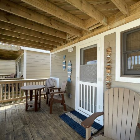 Wooden porch with two chairs, a small round table, a doormat, and hanging fish-shaped wall decor by a screened door.