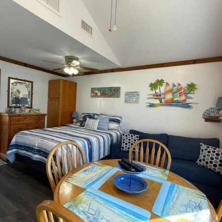 Cozy beach-themed room with a bed, sofa, dresser, ceiling fan, and dining table for four by a window.