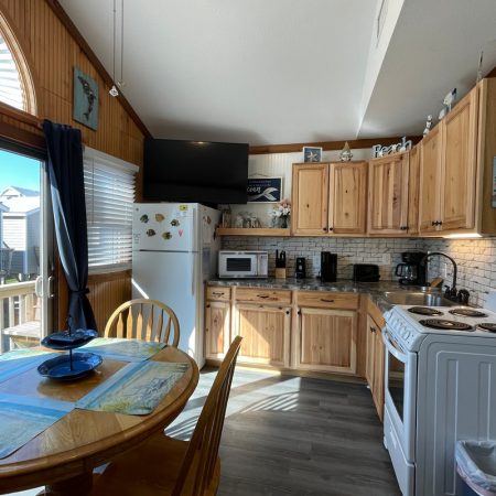 Small kitchen with wooden cabinets, dining table, white appliances, and sunlight coming through a large window.