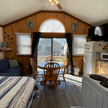 Small cabin interior with bed, sofa, dining table, kitchen, and large window facing a deck and neighboring houses.