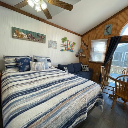 Bedroom with striped bedding, a blue couch, small dining table, and large window with a view of neighboring houses.