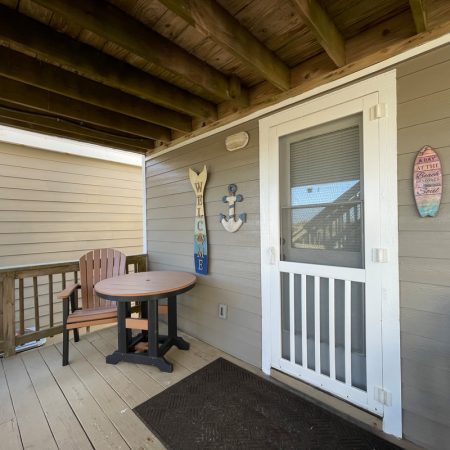 Small porch with two chairs, a round table, a screen door, and beach-themed wall decor including a “Welcome” sign.