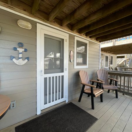 Porch with two chairs, a mat, wall decor, and a door under a wooden deck; stairs and railing in background.