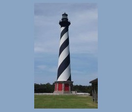 Tall lighthouse with black and white spiral stripes, standing on a grassy area with a clear sky in the background.