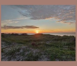 Sunset over a grassy dune with clouds in the sky and calm water in the distance.