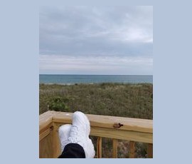 Person with white sneakers resting feet on a wooden railing, overlooking grassy dunes and the ocean under a cloudy sky.