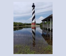 A black and white striped lighthouse stands by a flooded area, reflecting in the water next to a wooden pavilion.