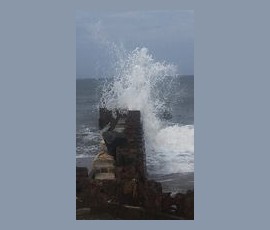 Ocean waves crash against a stone seawall under a cloudy sky, splashing water high into the air.