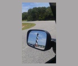 A black-and-white striped lighthouse is reflected in a car’s side mirror on a clear day, with trees in the background.