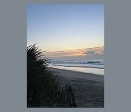 Ocean waves roll onto a sandy beach at sunrise, with grass and a fence in the foreground.