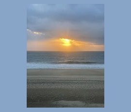 Sunset over a calm ocean with gentle waves, cloudy sky, and empty sandy beach in the foreground.