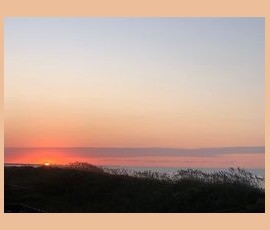 Sunset over a grassy dune with the sun partially visible on the horizon and a clear sky above.