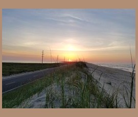 Sunrise over a sandy beach with tall grass, a road, and the ocean visible under a clear sky.