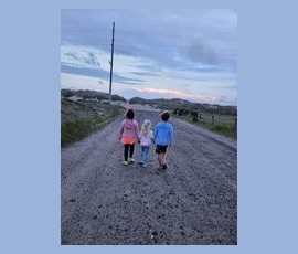 Three children walk down a gravel road at dusk, holding hands, with open fields and distant hills in the background.