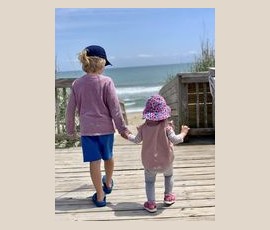 Two young children holding hands, walking on a wooden path toward a sandy beach and the ocean under a blue sky.
