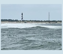 Choppy ocean waves with a striped lighthouse and a tall tower visible on the distant sandy shore under a cloudy sky.