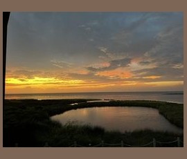 Sunset over a coastal landscape with a pond in the foreground and clouds illuminated by the setting sun.