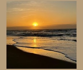 Sunrise over the ocean with waves gently washing onto a sandy beach under a clear sky.