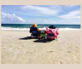 Two people sit on beach chairs facing the ocean, with sand, waves, and a partly cloudy sky in the background.
