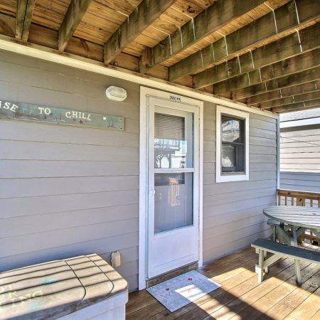 Wood deck with a white door, picnic table, storage box, and a “License to Chill” sign on gray siding under a wooden ceiling.