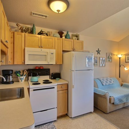 Small kitchen with light wood cabinets, white appliances, and a cozy living area with a loveseat and decor on the wall.