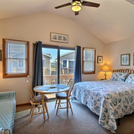 Bright bedroom with a bed, small dining table, sofa, TV, lamps, and windows leading to a small balcony.