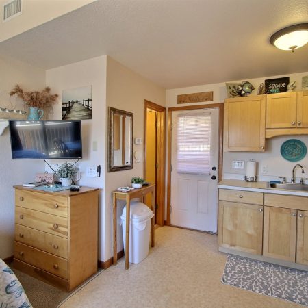 Small kitchenette with light wood cabinets, a sink, stove, dresser, TV, and beach-themed decor in a compact room.