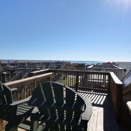 Two wooden chairs on a sunny rooftop deck overlook rooftops and the distant ocean under a clear blue sky.
