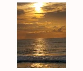 Golden sun sets over a calm ocean with gentle waves and clouds in the sky.
