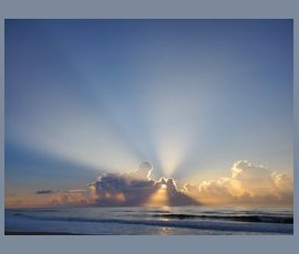 Sun rays burst through clouds over the ocean at sunrise, casting light on the water and sky.