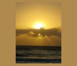 Sunset over the ocean with dark clouds partially covering the sun and gentle waves in the foreground.