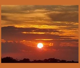 The sun sets behind dark clouds with an orange and yellow sky above a silhouetted treeline.