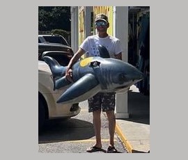 A person stands outside holding a large inflatable shark next to a parked white car on a sunny day.