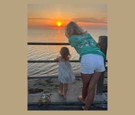 An adult and a child stand by a railing, watching the sun set over a calm ocean. A toy sits on the wooden deck.