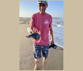 Man standing on a sandy beach holds a small fish in one hand and a device in the other, with waves in the background.