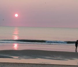 Person walks along the beach at sunset with calm waves and pink-purple sky reflecting on the water.