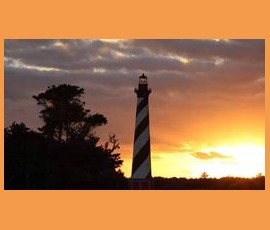 Striped lighthouse silhouetted against an orange sunset sky with trees in the foreground.