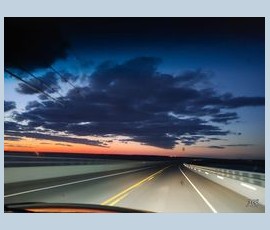 A view from a car driving on an empty road at sunset, with dark clouds and orange light on the horizon.