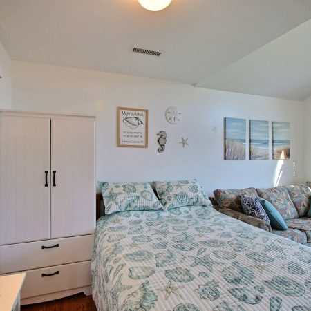 Bedroom with a bed, patterned bedding, a white wardrobe, a sofa, wall art, and two windows letting in natural light.