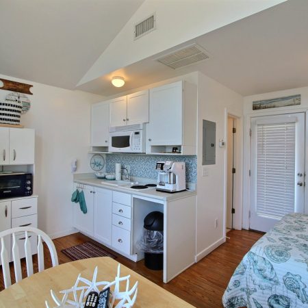 Compact studio with a bed, kitchenette, small dining area, and coastal decor. White cabinets and wood flooring.
