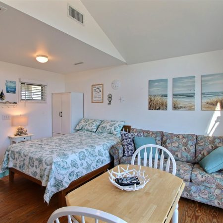 Bright studio with a bed, sofa, dining table, and beach-themed decor under natural and ceiling light.